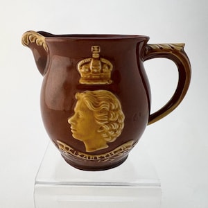May include: Brown ceramic pitcher with a gold-colored profile of a person and a crown. The pitcher has a handle and spout with gold accents. The design is embossed on the front of the pitcher.