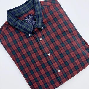 May include: A red and green plaid button-down shirt with a contrasting green and navy blue plaid collar. The shirt has white buttons and a label that reads "RUGBY" and "RALPH LAUREN".
