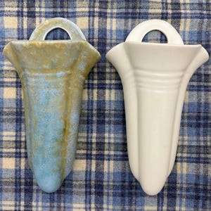 May include: Two ceramic wall vases. One vase is glazed in shades of blue and green, the other is white. Both vases have a flared top and a loop for hanging. The vases are displayed on a blue and white plaid fabric.
