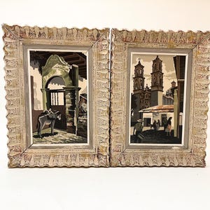 May include: Two framed paintings with ornate white frames. The painting on the left depicts a donkey standing in a doorway with a green archway. The painting on the right depicts a church with two towers and people walking in the street.