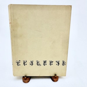 May include: A vintage book with a light tan cover. The cover features a row of small black cartoon figures along the bottom edge. The book is propped up on a wooden stand, showcasing its aged appearance and design.