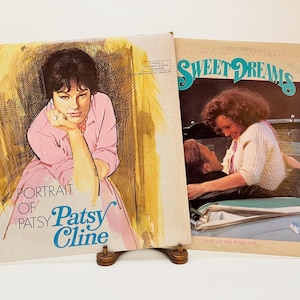 May include: Two vinyl record albums. The album on the left is a portrait of Patsy Cline with the title "Portrait of Patsy Cline". The album on the right is a portrait of a couple in a car with the title "Sweet Dreams: The Life and Times of Patsy Cline".