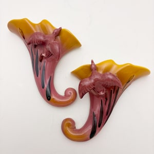 May include: Pair of vintage wall sconces in a horn shape, featuring a bird figurine. The sconces are painted in shades of pink, orange, and yellow, with black accents. These decorative pieces are likely made of ceramic or a similar material.