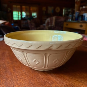 May include: A round ceramic mixing bowl with a pale yellow interior and a cream-colored exterior. The outer surface features a decorative embossed pattern consisting of a zigzag border along the rim and a series of rectangular panels with diamonds.