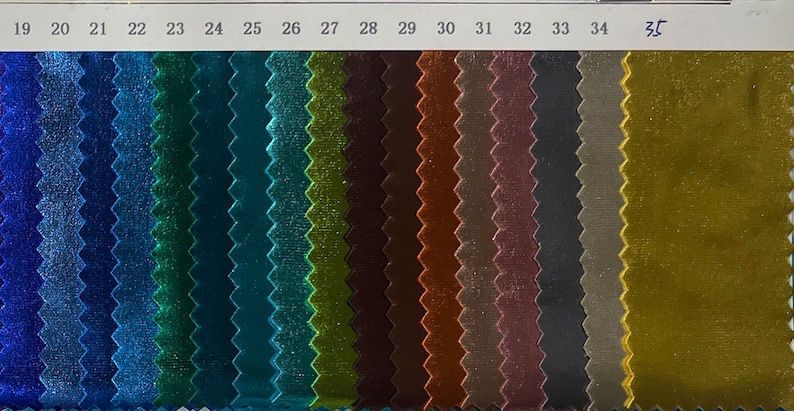 May include: A colour swatch card featuring samples of shiny fabric in various colours. The samples are arranged in vertical rows, showcasing shades of blue, green, brown, and gold. The numbers 19 to 35 are printed at the top.