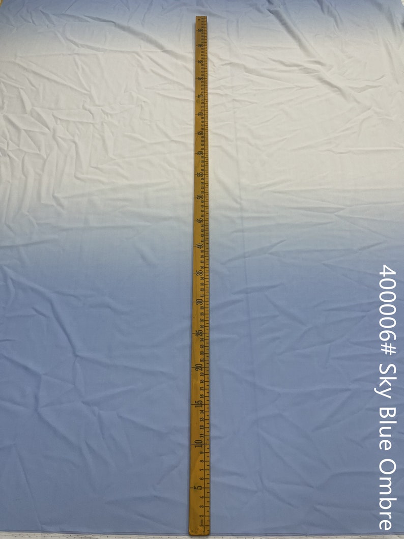 May include: A close-up of a light blue ombre fabric with a wooden ruler measuring 40 inches. The fabric is smooth and has a slight sheen. The ruler is marked in inches and has a metal tip.