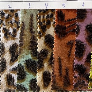 May include: A fabric swatch card displaying seven animal print designs. The patterns, reminiscent of leopard and cheetah, feature shades of brown, black, orange, and yellow. The fabric has a textured finish.