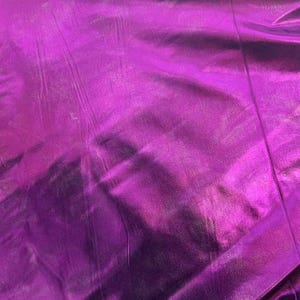 May include: A close-up shot of a vibrant, metallic purple fabric. The material has a reflective surface with subtle folds and creases, creating highlights and shadows. The overall texture appears smooth and luxurious, suitable for clothing or decorative projects.
