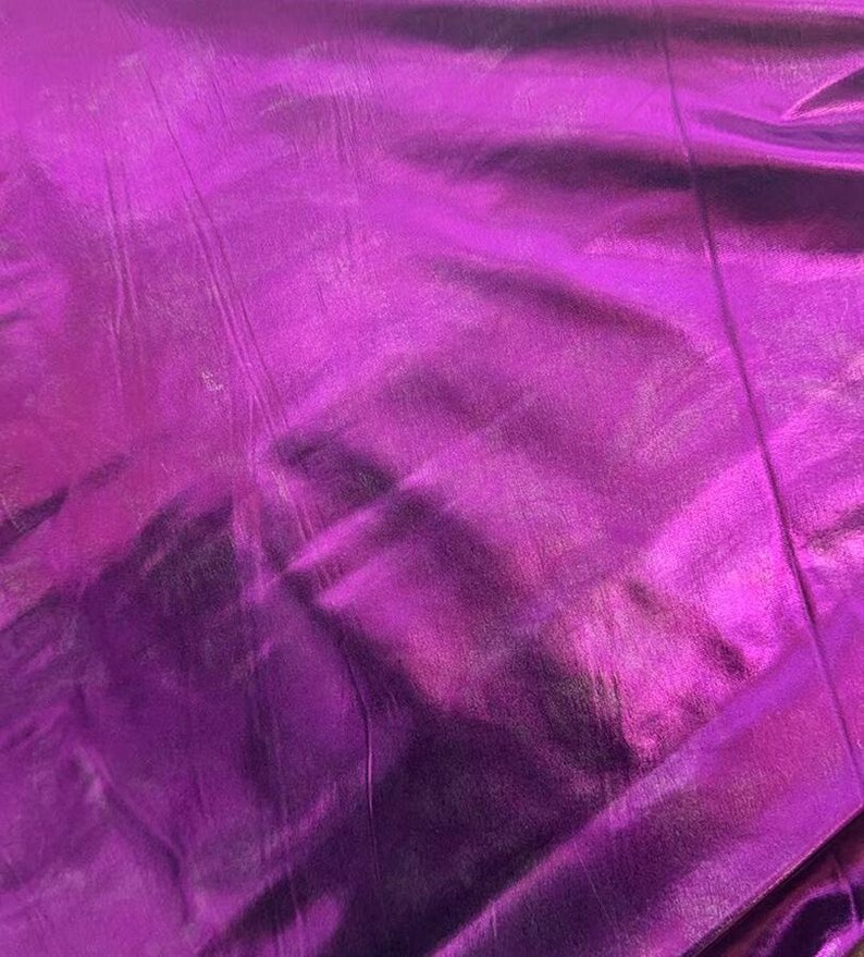 May include: A close-up shot of a vibrant, metallic purple fabric. The material has a reflective surface with subtle folds and creases, creating highlights and shadows. The overall texture appears smooth and luxurious, suitable for clothing or decorative projects.