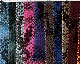 May include: A selection of fabric samples featuring a snakeskin design in diverse colours. The swatches showcase hues of blue, green, pink, purple, brown, and black. The fabric has a textured finish, and the edges are cut in a zig-zag pattern.