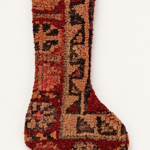 May include: A holiday stocking crafted from a textured, looped yarn material. The surface features a geometric pattern in deep shades of red, burnt orange, brown, and tan. The design includes repeating square motifs and linear borders throughout.