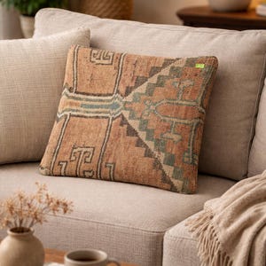 Peut inclure: Coussin carré avec un motif géométrique dans les tons marron, beige et sarcelle. Le coussin est posé sur un canapé beige. Une table basse avec une tasse de café et un livre est au premier plan.
