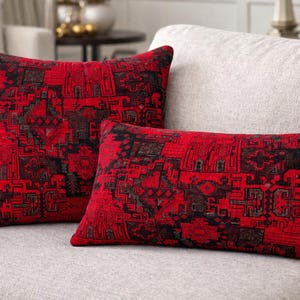 Peut inclure: Deux coussins décoratifs avec un motif géométrique rouge et noir. L'un est carré, l'autre rectangulaire. Les coussins sont sur un canapé gris clair, ajoutant une touche de couleur à l'ensemble neutre.