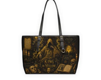 May include: A black tote bag featuring a gold-toned illustration of a hooded figure, a skull, and open books. The text reads The King in Yellow, along with smaller passages. The bag has two black shoulder straps with silver-toned metal buckles.