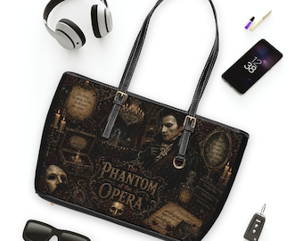 May include: A black tote bag featuring a detailed graphic print of The Phantom of the Opera, including a portrait of the Phantom, a white mask, a chandelier, and text. The bag has two black shoulder straps with gold-toned buckles and metal hardware.