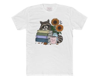 May include: A white t-shirt featuring a graphic illustration of a raccoon resting on a stack of three books. Next to the books are two sunflowers in a glass jar, a pink mug with the text Wise Hazel, and a raccoon tail curled near the base.