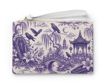 May include: A rectangular wristlet clutch featuring a purple and cream toile-style print. The design depicts ravens, coffins, skulls, lilies, a bridge, a pagoda, and a crescent moon. It includes a white wrist strap and a gold-toned zipper closure.