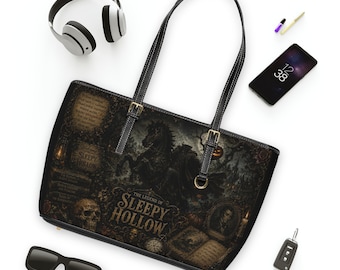 May include: A black tote bag featuring a dark, gothic-style illustration of The Legend of Sleepy Hollow, including a horse, a skull, and text. The bag has two black shoulder straps with gold-tone buckles and a structured rectangular shape.