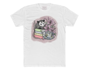 May include: A white short-sleeve t-shirt featuring a graphic illustration of a black and white opossum perched on a stack of four pastel-colored books next to a floral arrangement of pink lilies and a teacup with a tea bag tag labeled Tea Time.