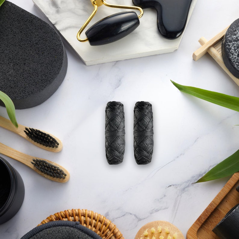 May include: Two black charcoal floss picks on a white marble surface. The picks are wrapped in black paper and have a woven texture.