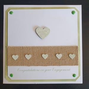 May include: A handmade engagement card with a white background, a burlap ribbon, and decorative hearts. The card features the text "Congratulations on your Engagement" in a script font. Green accents and embellishments complete the design.