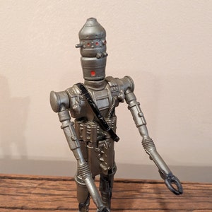 Peut inclure: Une figurine d'action en argent et noir d'un robot avec une t&ecirc;te en forme de d&ocirc;me et des yeux rouges. Le robot a une sangle noire sur la poitrine et tient ses bras tendus.