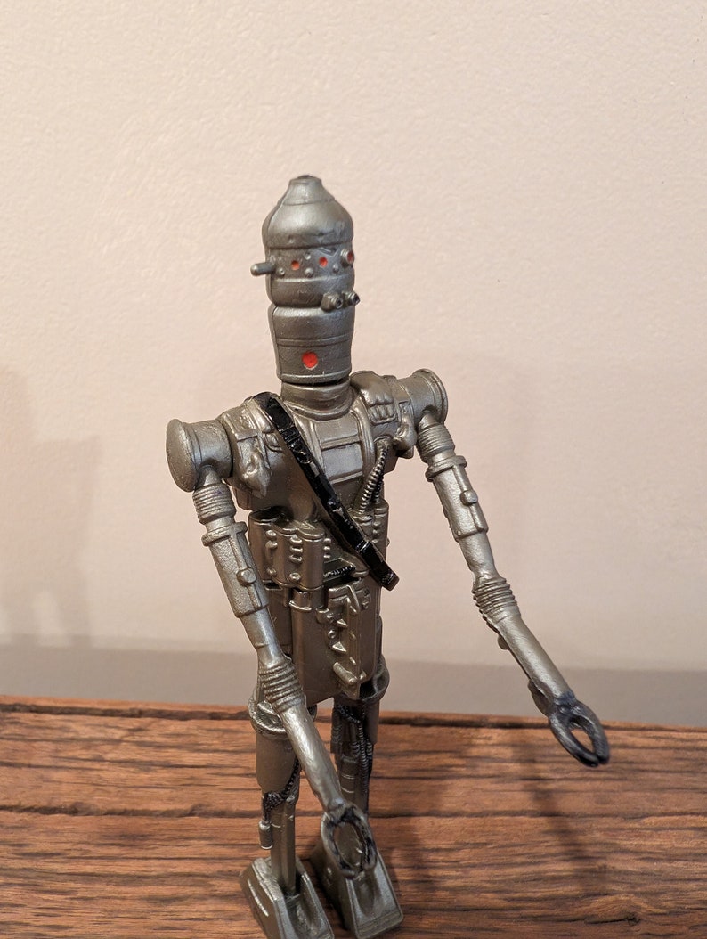 Peut inclure: Une figurine d'action en argent et noir d'un robot avec une t&ecirc;te en forme de d&ocirc;me et des yeux rouges. Le robot a une sangle noire sur la poitrine et tient ses bras tendus.