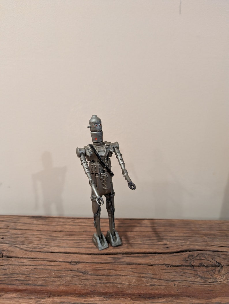 Peut inclure: Une figurine d'action en argent d'un robot avec un design d&eacute;taill&eacute;. Le robot a un corps long et fin avec une grosse t&ecirc;te et un petit torse rond. Il a deux bras avec des mains ouvertes et deux jambes avec des pieds plats sur le sol. Le robot est debout sur une surface en bois.