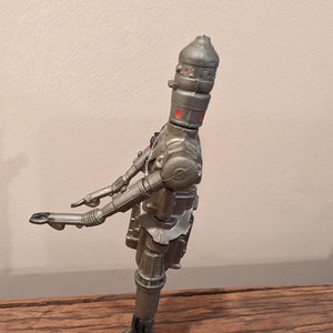 Peut inclure: Un jouet robot de couleur argent avec des yeux rouges et une t&ecirc;te en forme de d&ocirc;me. Le robot est debout sur une surface en bois.
