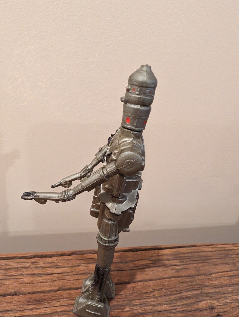 Peut inclure: Un jouet robot de couleur argent avec des yeux rouges et une t&ecirc;te en forme de d&ocirc;me. Le robot est debout sur une surface en bois.