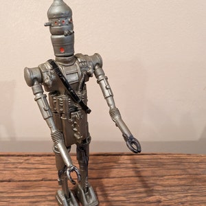 Peut inclure: Une figurine d'action de robot argent&eacute;e et noire avec un design d&eacute;taill&eacute;. Le robot a une t&ecirc;te en forme de d&ocirc;me avec des yeux rouges et une sangle noire sur la poitrine.