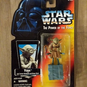 May include: A Star Wars action figure of Yoda, a green alien with large ears, wearing a brown robe and holding a stick. The figure is packaged in a plastic blister pack with a black and yellow background. The packaging features the Star Wars logo and the text "The Power of the Force".