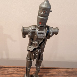 Peut inclure: Une figurine d'action en argent d'un robot avec une t&ecirc;te cylindrique et de multiples articulations. Le robot a une sangle noire sur la poitrine et se tient debout sur une surface en bois.