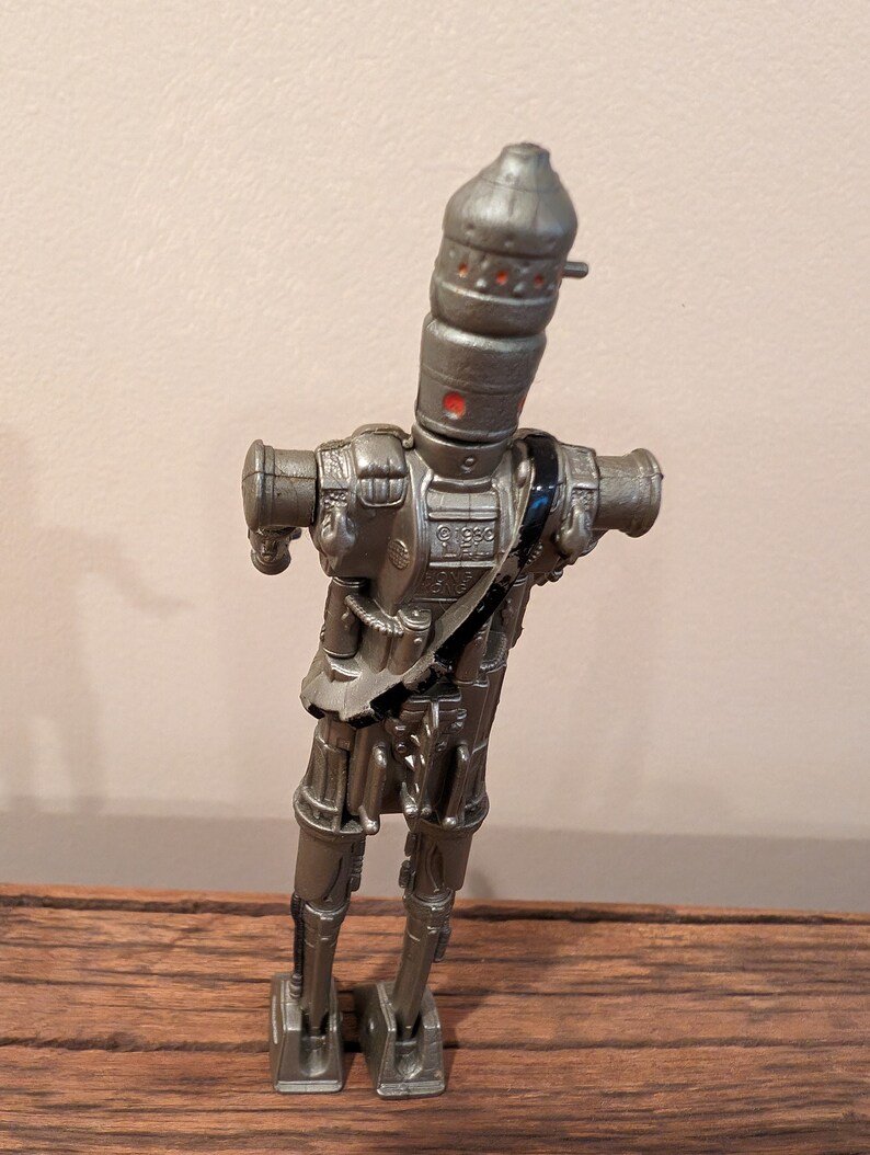 Peut inclure: Une figurine d'action en argent d'un robot avec une t&ecirc;te cylindrique et de multiples articulations. Le robot a une sangle noire sur la poitrine et se tient debout sur une surface en bois.