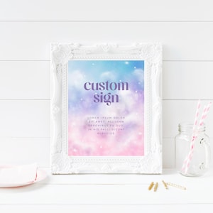 May include: A white framed sign with the words "custom sign" in purple lettering. The background of the sign is a dreamy blend of blue, pink, and purple hues. A pink plate, a glass jar with straws, and gold paper clips are also in the image.