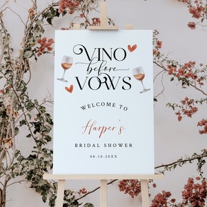 Pode incluir: Uma placa branca com o texto "Vino before vows" em escrita cursiva, com duas ta&ccedil;as de vinho e cora&ccedil;&otilde;es. A placa tamb&eacute;m diz "Bem-vindo ao Bridal Shower de Harper 08.10.20XX". A placa est&aacute; em um cavalete de madeira.