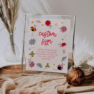 May include: A printable sign with a pink background and a white frame. The sign features a custom text that says "custom sign" in red cursive font. The sign is decorated with red ladybugs, blue flowers, and green shamrocks. The text below the sign says "LOREM IPSUM DOLOR SIT AMET, ACCUSAM ERRORIBUS EU DUO, IN HIS FALLI DICUNT ALBUCIUS."