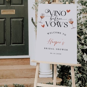 Pode incluir: Um cartaz branco com texto preto que diz "Vino before Vows" com um cora&ccedil;&atilde;o vermelho e ilustra&ccedil;&otilde;es de ta&ccedil;as de vinho. O cartaz tamb&eacute;m diz "Welcome to Harper's Bridal Shower 08.10.20XX". O cartaz est&aacute; em um cavalete de madeira.