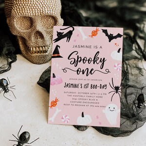May include: A pink Halloween party invitation featuring a black cat, witch's hat, bats, sweets, and pumpkins. The text reads "Jasmine is a Spooky One" and "Jasmine's 1st Boo-Day".