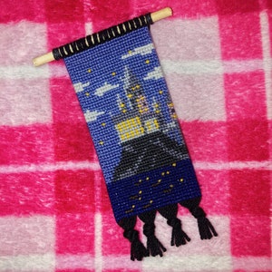 May include: A blue and black tapestry with a stitched image of a house with yellow windows and a dark blue sky with white clouds and yellow stars. The tapestry has black tassels at the bottom.