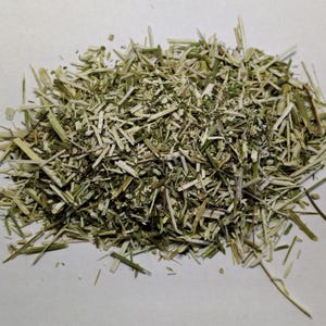 May include: A pile of dried, chopped herbs, primarily green and tan in color. The pieces vary in size and shape, with some appearing like small stems or leaves. The herbs are scattered on a light-colored surface.