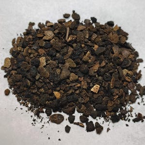 May include: A close-up shot of a pile of dark brown, irregularly shaped pieces, possibly bark or wood chips, on a white surface. The pieces vary in size and texture, with some lighter brown fragments mixed in.
