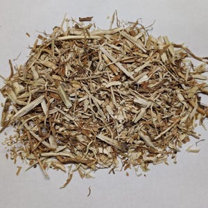 May include: A pile of dried, light brown plant material, possibly herbs or roots, with a variety of shapes and textures. The pieces vary in size, from thin strands to small chunks, and are scattered across a white surface.