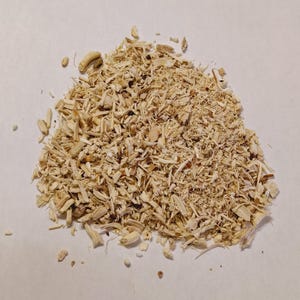 May include: A pile of light beige, shredded wood chips. The chips vary in size and shape, with some longer and thinner than others. The background is a plain, off-white surface.