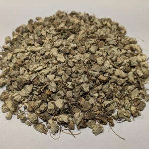 May include: A pile of small, irregularly shaped, light brown pieces, possibly herbs or spices, with some visible stems and fibers. The pieces have a rough, textured appearance, suggesting they are dried or processed.