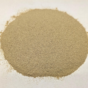 May include: A circular pile of fine, light brown powder, possibly a spice or ingredient, sits on a white surface. The powder has a slightly coarse texture and is evenly distributed, with a few grains scattered around the edges.