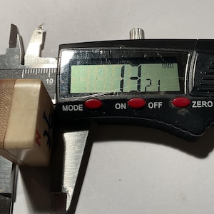 May include: A rectangular mahjong tile with a cream-colored face and a brown bamboo backing, positioned within a black digital caliper. The digital display clearly shows a measurement of 14.21 mm, indicating the thickness of the tile piece.