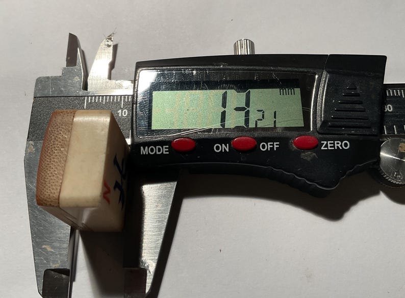 May include: A rectangular mahjong tile with a cream-colored face and a brown bamboo backing, positioned within a black digital caliper. The digital display clearly shows a measurement of 14.21 mm, indicating the thickness of the tile piece.