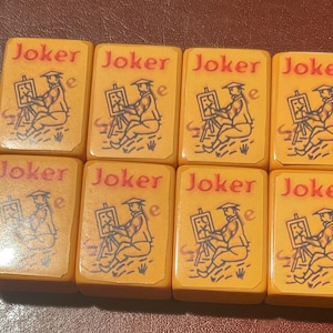 Puede incluir: Ocho fichas de Mahjong vintage. Cada ficha es rectangular con esquinas redondeadas y un fondo amarillo-naranja. La palabra "Joker" est&aacute; en rojo en la parte superior. Una figura sentada pintando en un caballete est&aacute; representada en negro.