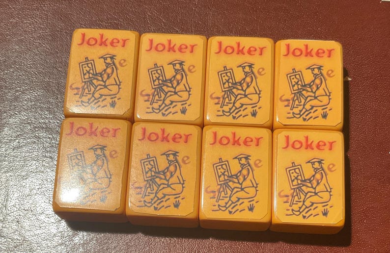 Puede incluir: Ocho fichas de Mahjong vintage. Cada ficha es rectangular con esquinas redondeadas y un fondo amarillo-naranja. La palabra "Joker" est&aacute; en rojo en la parte superior. Una figura sentada pintando en un caballete est&aacute; representada en negro.
